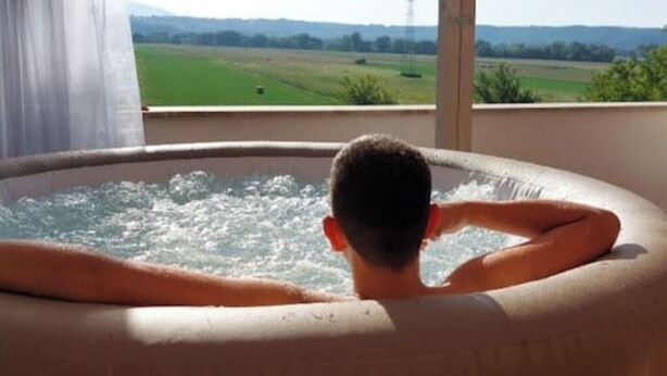 Una persona relajándose en un jacuzzi con vista a un campo verde y una torre a lo lejos.