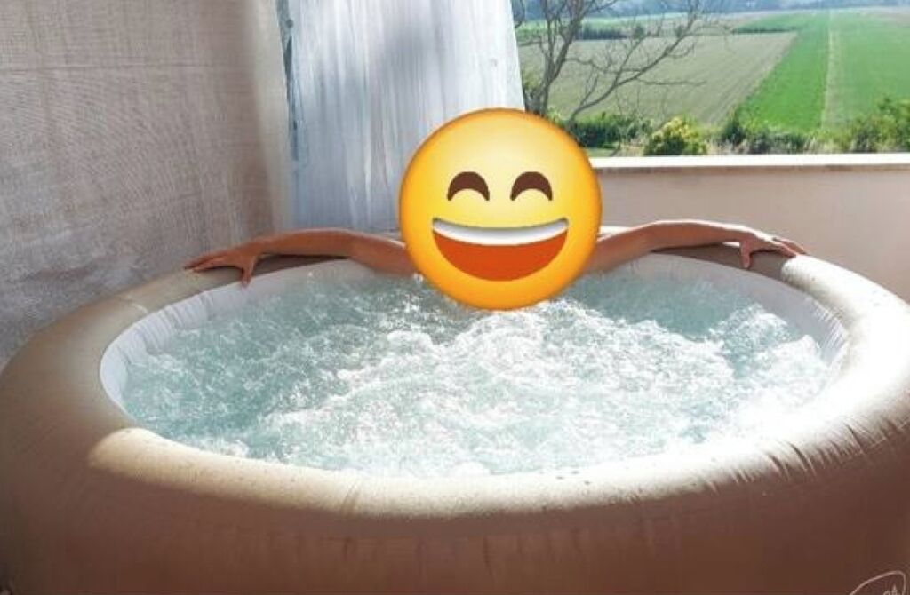 Una vasca idromassaggio con un'emoji sorridente al centro, circondata da una finestra che offre vista su un paesaggio verde.