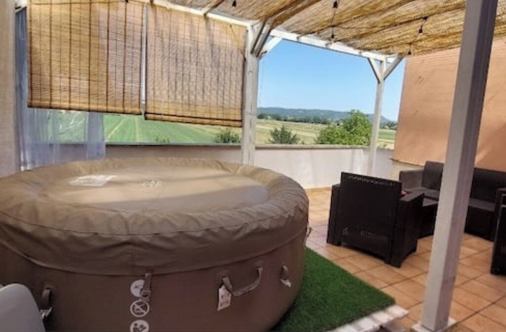 Ein Whirlpool mit Blick auf eine grüne Landschaft, eine überdachte Terrasse mit Tisch und Stühlen und ein Strohdach.