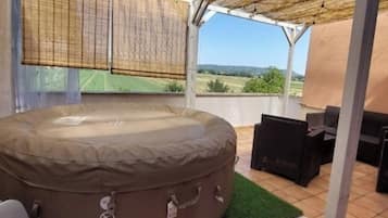 Ein Whirlpool mit Blick auf eine grüne Landschaft, eine überdachte Terrasse mit Tisch und Stühlen und ein Strohdach.