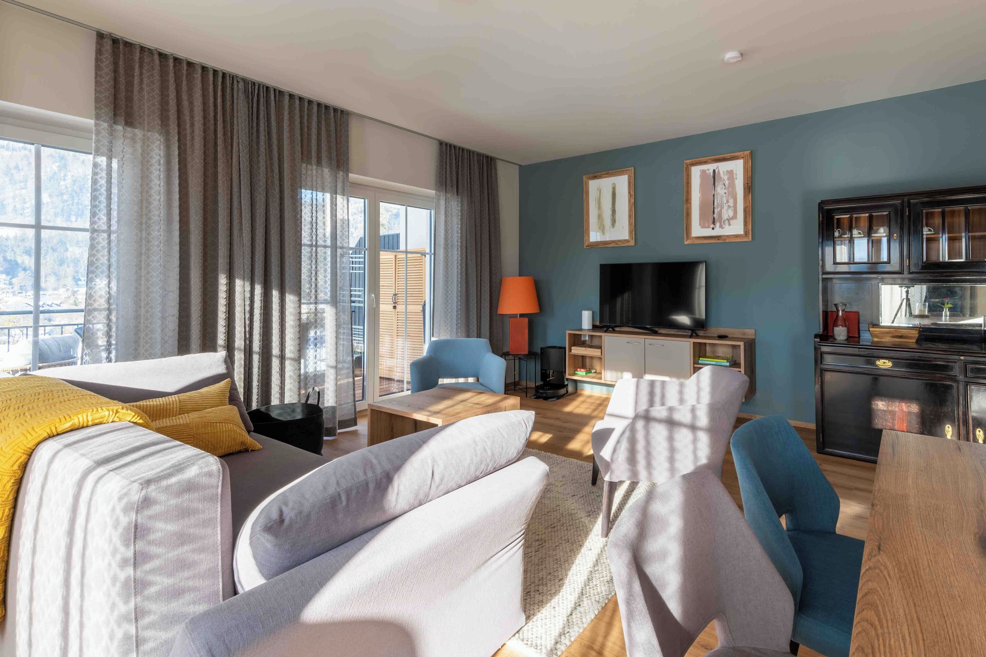 A modern hotel room with a sofa, armchairs, a dining table, a TV, and a kitchen area.