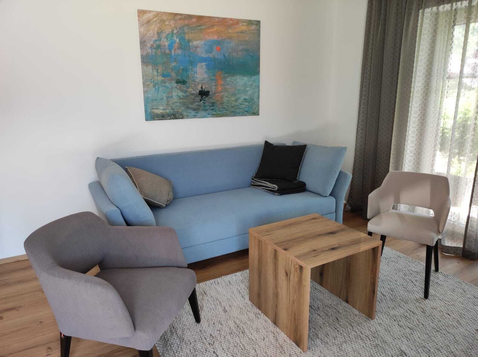 Un salon avec un canapé bleu, un fauteuil gris, une table basse en bois et un tableau accroché au mur.