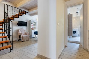A staircase with wooden steps and black railings, a white wall with a mounted television, a chair with a striped cover, and a laundry area with a washing machine.