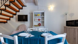 Un área de comedor con mantel azul, sillas blancas, una estantería y un televisor montado en la pared.