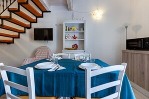 A dining area with a blue tablecloth, white chairs, a bookshelf, and a TV mounted on the wall.