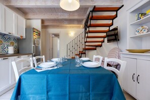 Un coin repas avec une nappe bleue, des chaises blanches, un escalier en bois et une cuisine avec des meubles blancs.