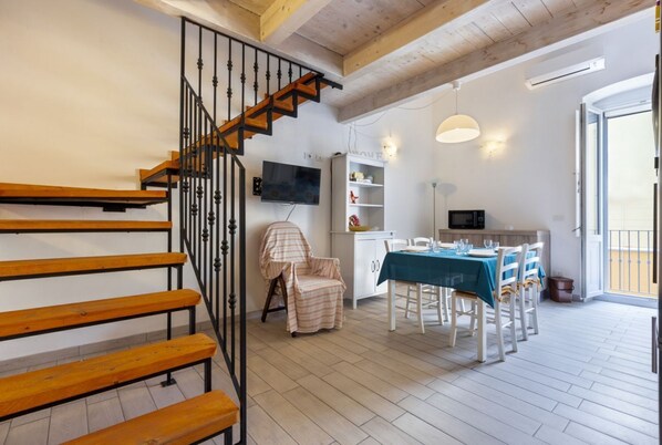 Un escalier avec des marches en bois et des rampes en métal, un coin repas avec une table dressée pour quatre personnes, un coin cuisine équipé d’un four à micro-ondes et un téléviseur fixé au mur.