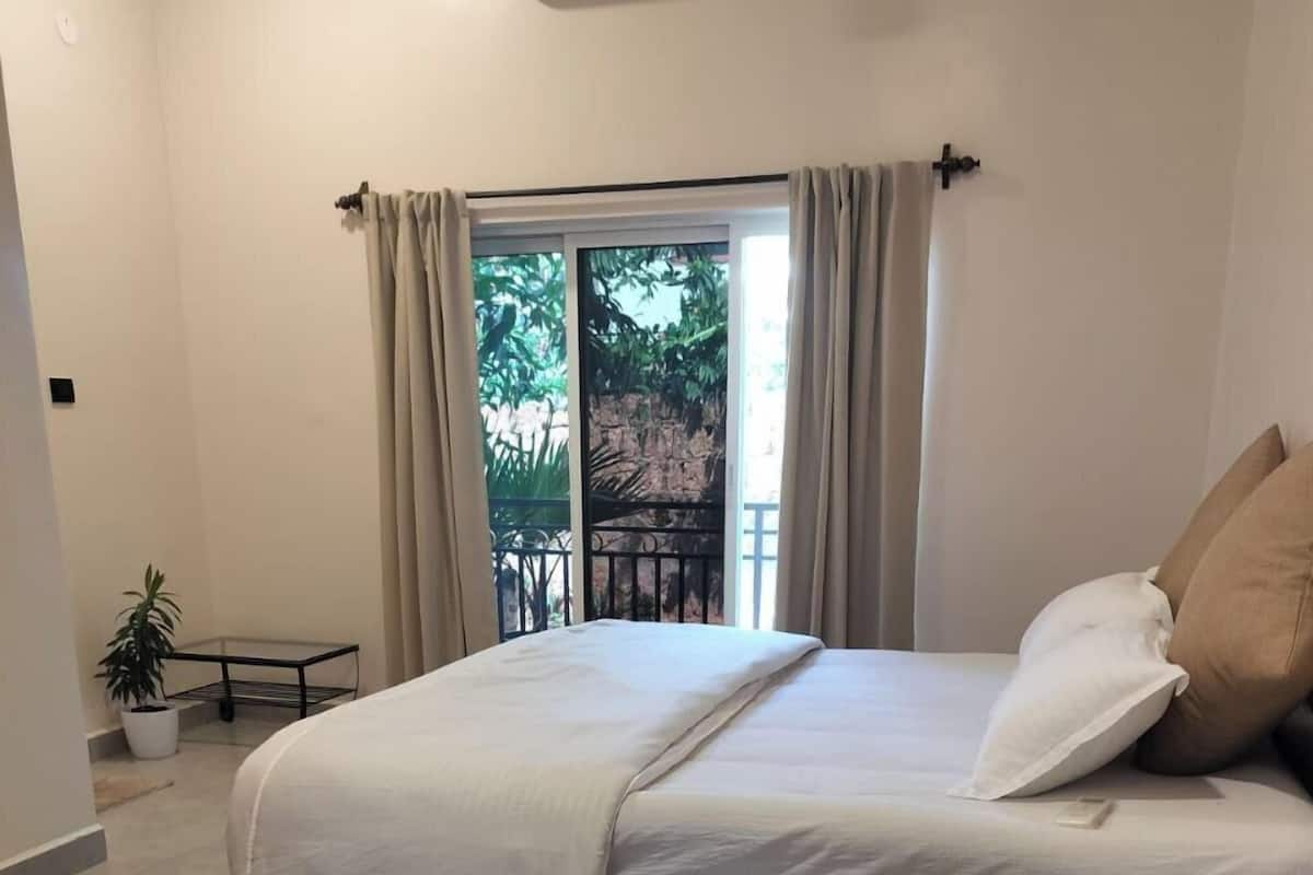 Quarto com cama, cortinas, um vaso com planta e vista para o verde lá fora.
