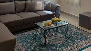 Een woonkamer met een bruine bank, een glazen bijzettafeltje met fruit, en een vloerkleed met een patroon.
