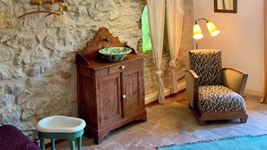 A rustic room with a stone wall, wooden beams, a wooden cabinet with a bowl, a green chair, a small stool, and a blue rug.
