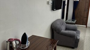 Ein Wohnzimmer mit grauem Sofa, einem Holztisch mit Wasserkocher und Bügeleisen, und einem an der Wand montierten Fernseher.