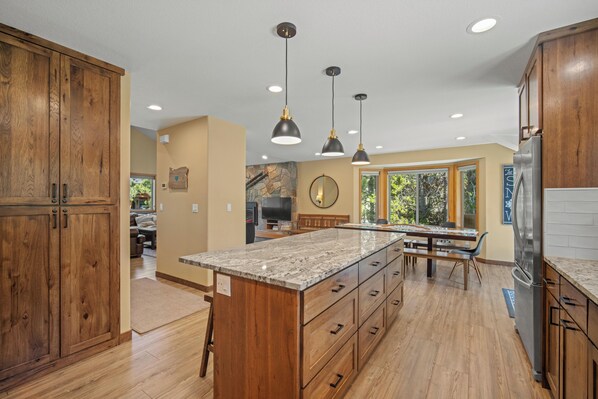 Una cocina amplia con armarios de madera, una isla de encimera de granito, iluminación colgante y un ventanal que proporciona luz natural.