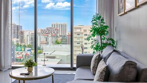 A modern living room with a large window, a grey sofa, a small round table, and a potted plant.