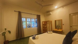 Habitación de hotel con cama, ventilador de techo, ventana con cortinas, una mesita con espejo y una planta en maceta.