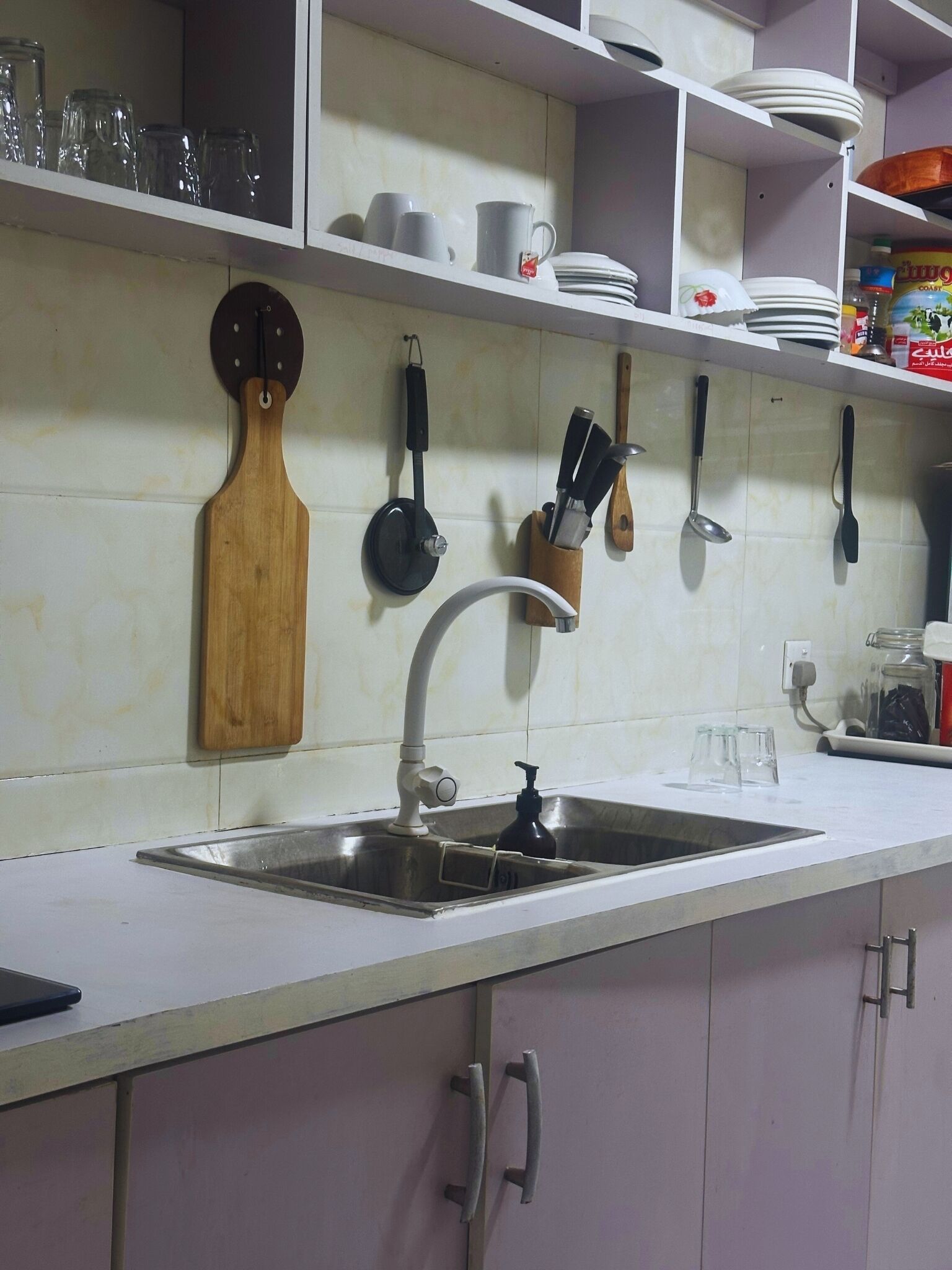 A kitchen with a stainless steel sink, wooden cutting board, hanging utensils, and shelves with dishes and jars.