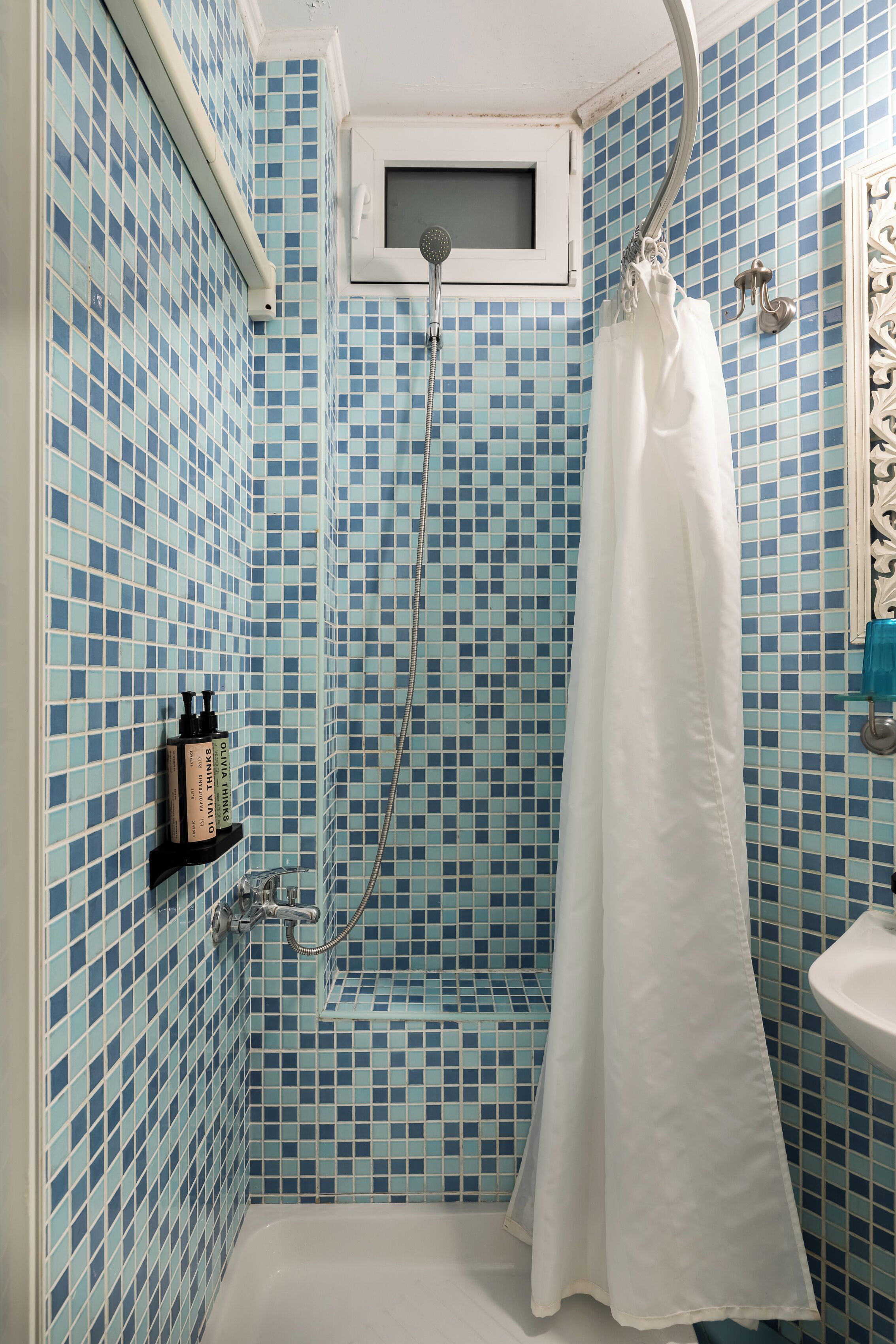 A bathroom with blue mosaic tiles, a white shower curtain, a handheld showerhead, and a sink with a blue soap dispenser.