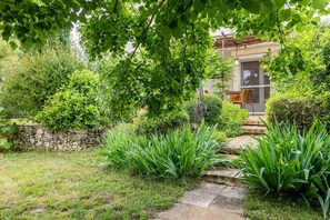 A garden with a stone wall, a pathway leading to a building with a door and a chair outside.