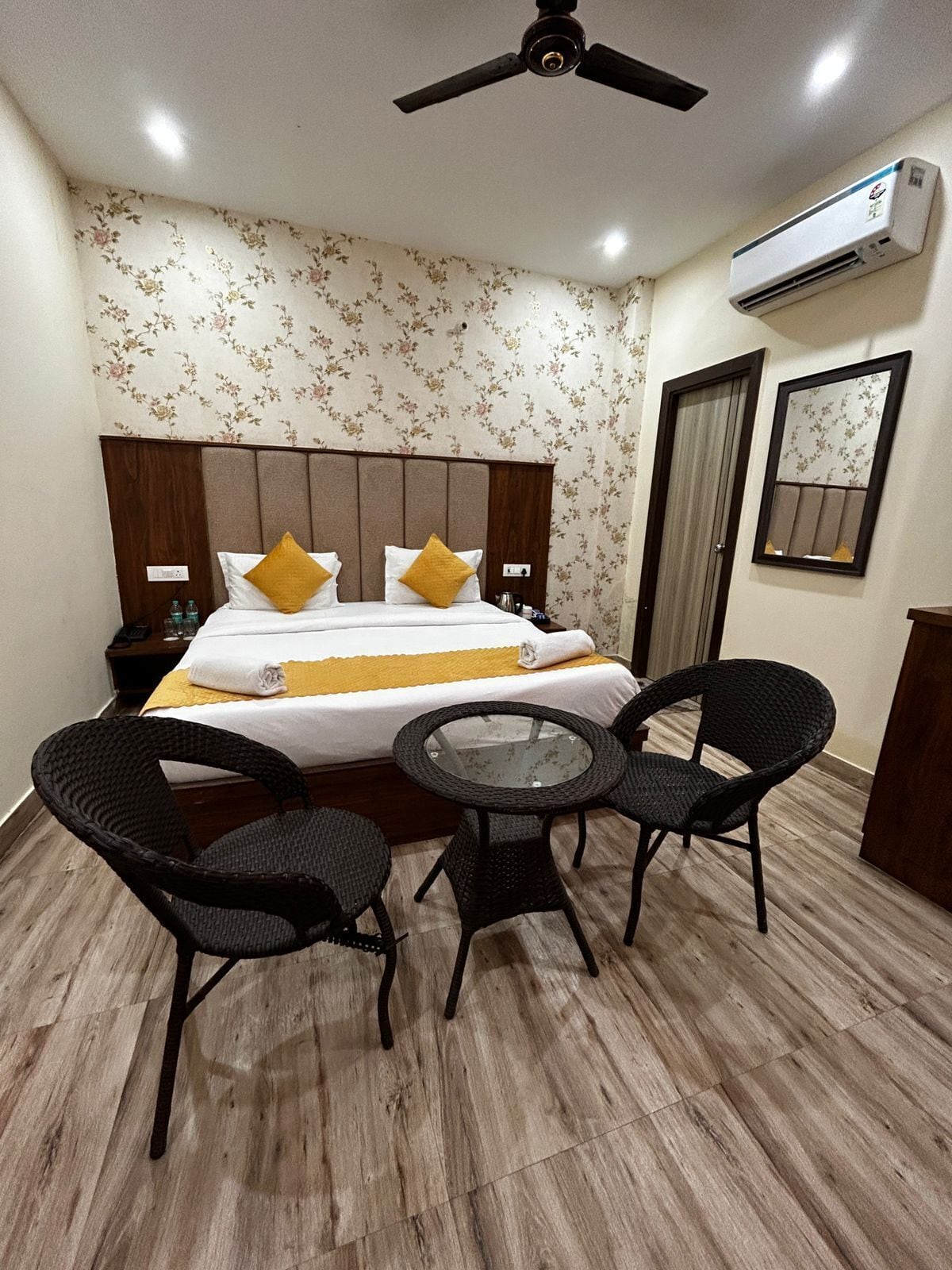 A hotel room with a bed, two chairs, a table, a ceiling fan, an air conditioning unit, and a mirror on the wall.