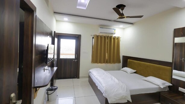 A hotel room with a bed, a ceiling fan, an air conditioning unit, and a door. - Hotel Vardhman (Ujjain)