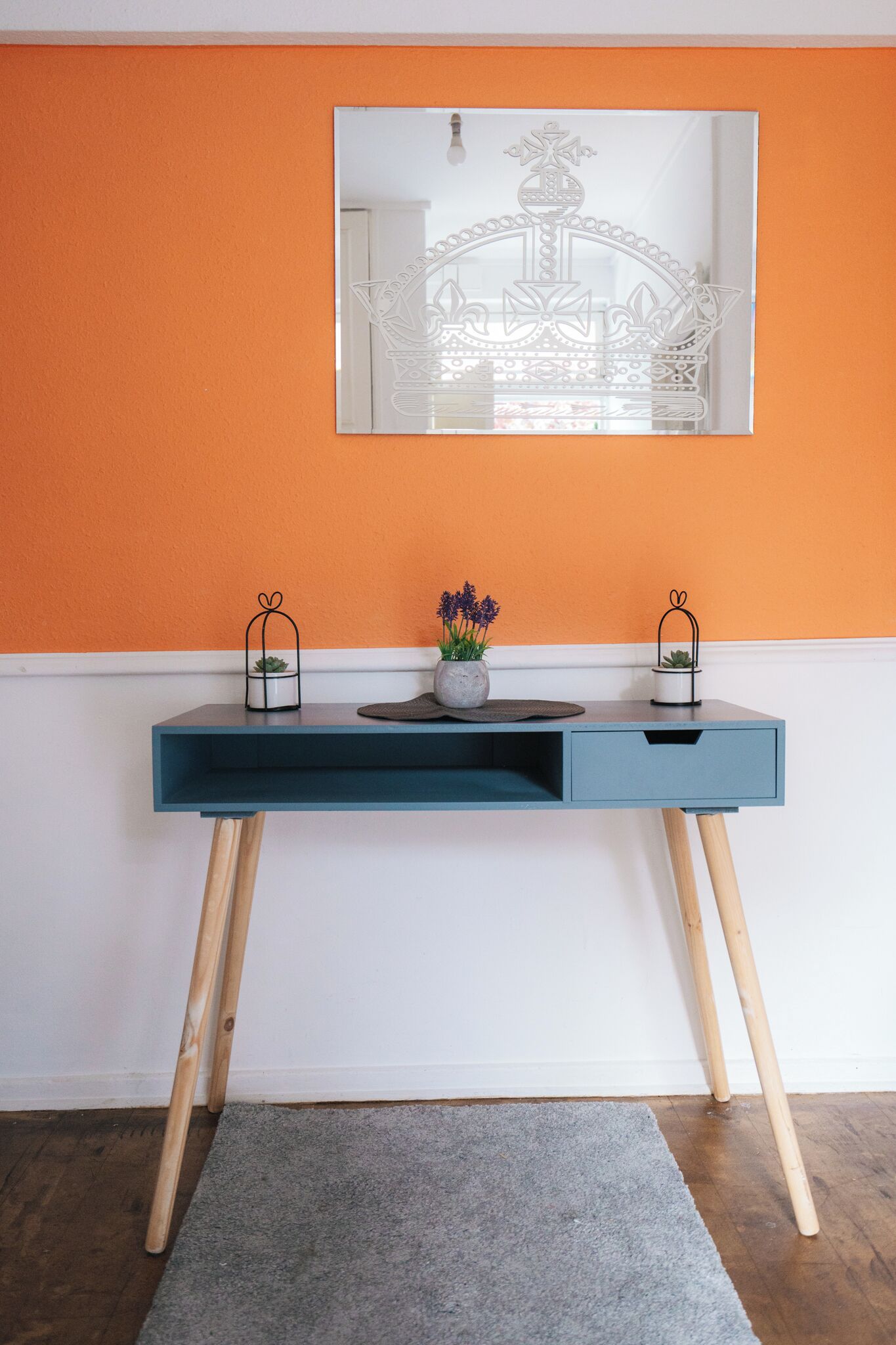 Een modern bureau met een spiegel, twee planten in potten en een grijs vloerkleed in een kamer met oranje en witte wanden.