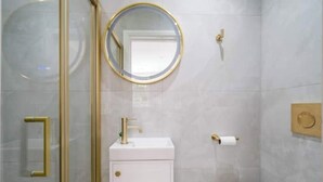 A bathroom with a glass shower enclosure, a white sink with gold fixtures, a round mirror, and a toilet. - Premium - 1 BR Flat with Terrace (Kentish Town)
