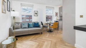 A modern living room with a sofa, a dining table, and chairs. There are windows with a view of buildings outside. - Premium - 1 BR Flat with Terrace (Kentish Town)