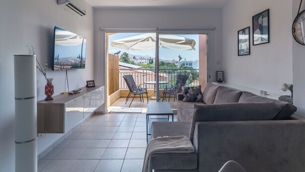 A modern living room with a grey sofa, a coffee table, and a TV on the wall. There is a sliding glass door leading to an outdoor patio with umbrellas and chairs. - RelaxInCyprus 1-bedroom apartment in awesome Pegeia with WiFi, AC (Pegeia)
