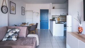 A modern living room with a sofa, a dining table, and chairs. A kitchen is visible in the background with white cabinets and a blue door. - RelaxInCyprus 1-bedroom apartment in awesome Pegeia with WiFi, AC (Pegeia)