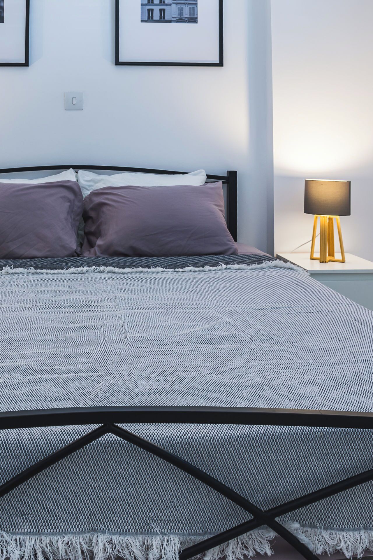 Una cama bien hecha con una colcha estampada y dos almohadas, una mesita de noche con una lámpara, y dos cuadros enmarcados en la pared.