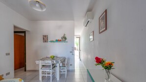 Une chambre d’hôtel avec un lit, une table à manger avec des chaises, un vase avec des fleurs, une télécommande et un couloir avec une porte et une étagère fixée au mur.