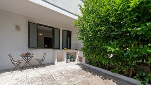Une terrasse avec une table et des chaises, un mur blanc et une haie verte.