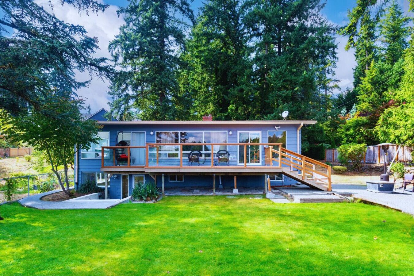 A modern two-story house with a deck, surrounded by lush greenery and tall trees.