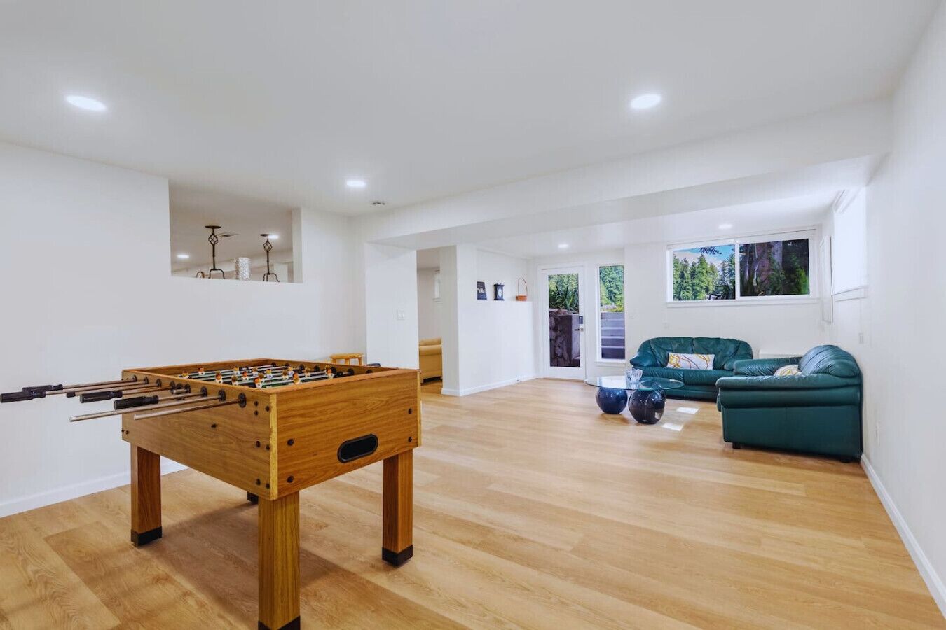 A spacious living room with a wooden foosball table, a green sofa, and a window with a view of greenery.