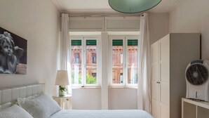 A bedroom with a bed, a nightstand, a lamp, a fan, and a view of a building through the window. - GuestHost - Cozy apartment of about 70 sqm, able to accommodate up to 4 people, located on the fourth floor of a building with a lift (NOT suitable for disabled people).The property is located in the Flaminio district, close to the Lungotevere, in a well (Rome)