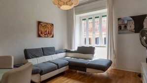 A modern living room with a sectional sofa, a painting, a window with a view of buildings, and a fan. - GuestHost - Cozy apartment of about 70 sqm, able to accommodate up to 4 people, located on the fourth floor of a building with a lift (NOT suitable for disabled people).The property is located in the Flaminio district, close to the Lungotevere, in a well (Rome)