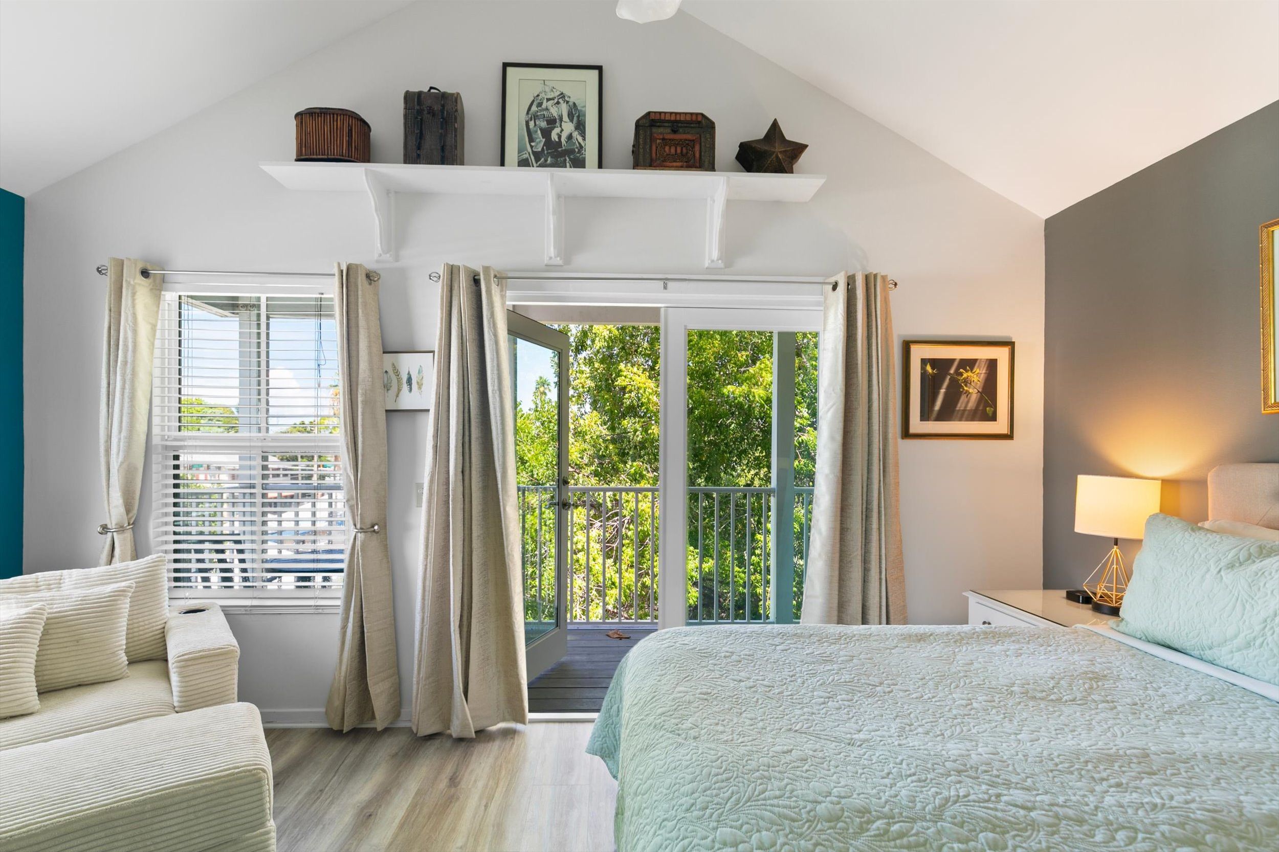 Quarto com cama, mesa de cabeceira, luminária, janela com cortinas e varanda com guarda-corpo.