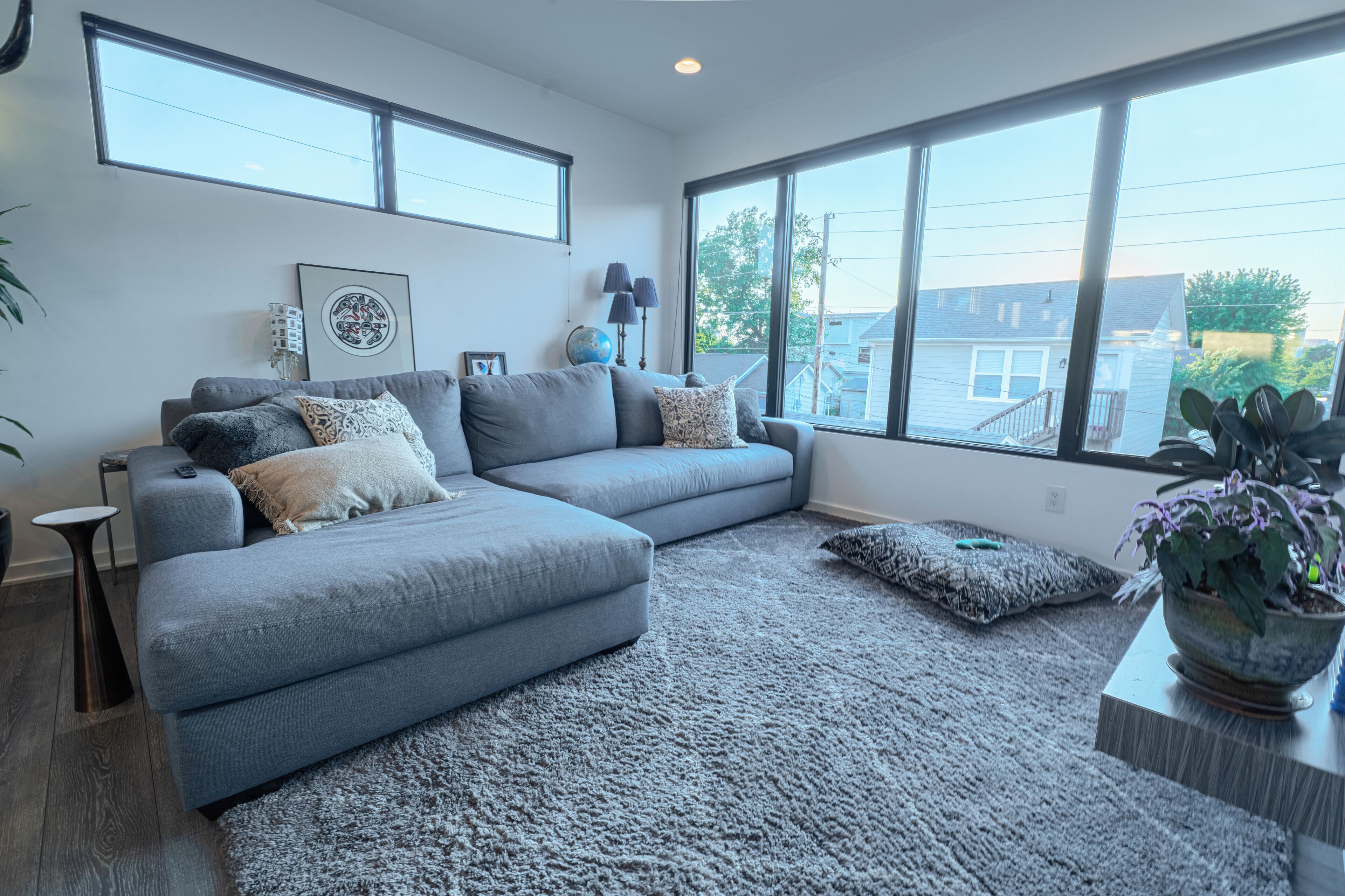 A modern living room with a grey sectional sofa, a large window with a view of the neighborhood, and a potted plant on a side table.