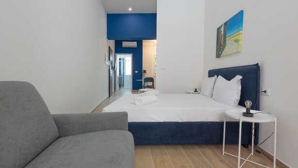 A modern hotel room with a large bed, a gray armchair, a small white table, and a painting on the wall. - Studio apartment with double bed, armchair bed and equipped kitchenette. (Turin)
