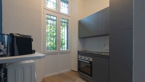 A compact kitchen with a built-in oven, a stove, and a refrigerator. There are two windows with a view of greenery outside. - Studio apartment with double bed, armchair bed and equipped kitchenette. (Turin)