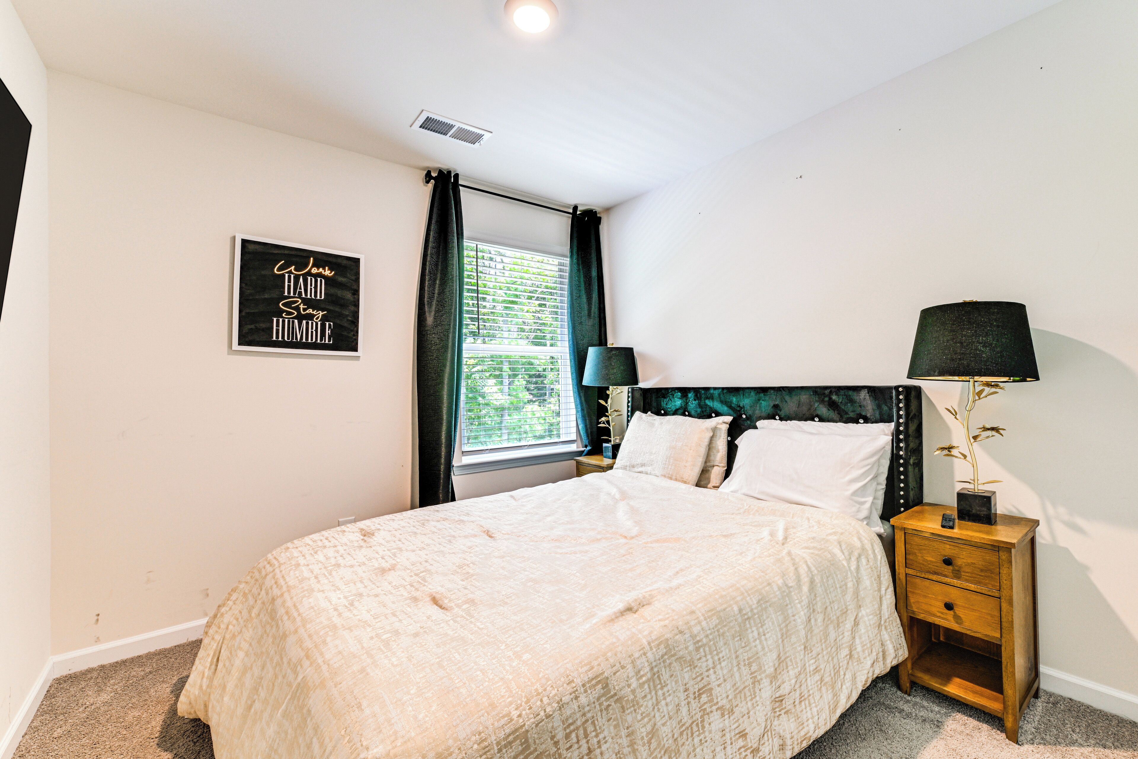A bedroom with a bed, a nightstand, a lamp, a window with curtains, and a motivational quote on the wall.