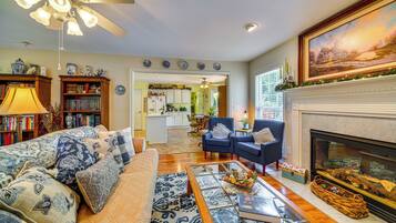 A living room with a sofa, armchairs, a fireplace, a bookshelf, and a kitchen in the background.