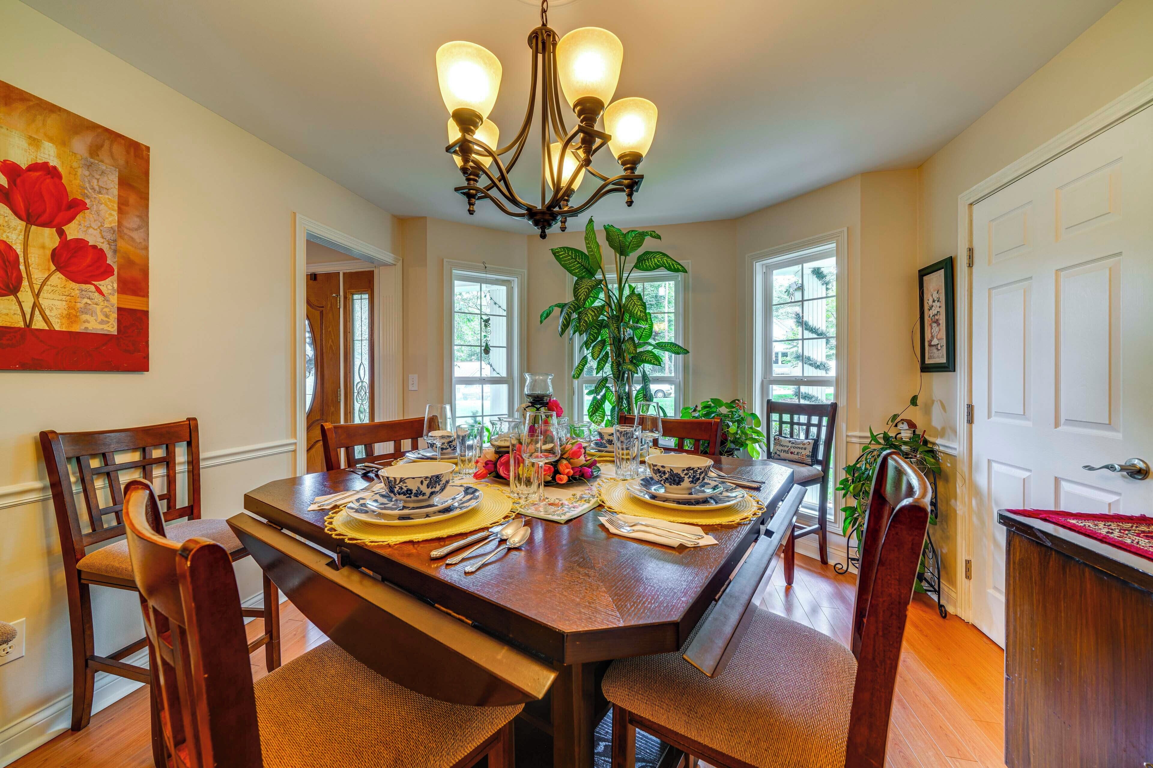 A dining room with a wooden table set for four, surrounded by chairs. A chandelier hangs from the ceiling, and there are windows on the walls.