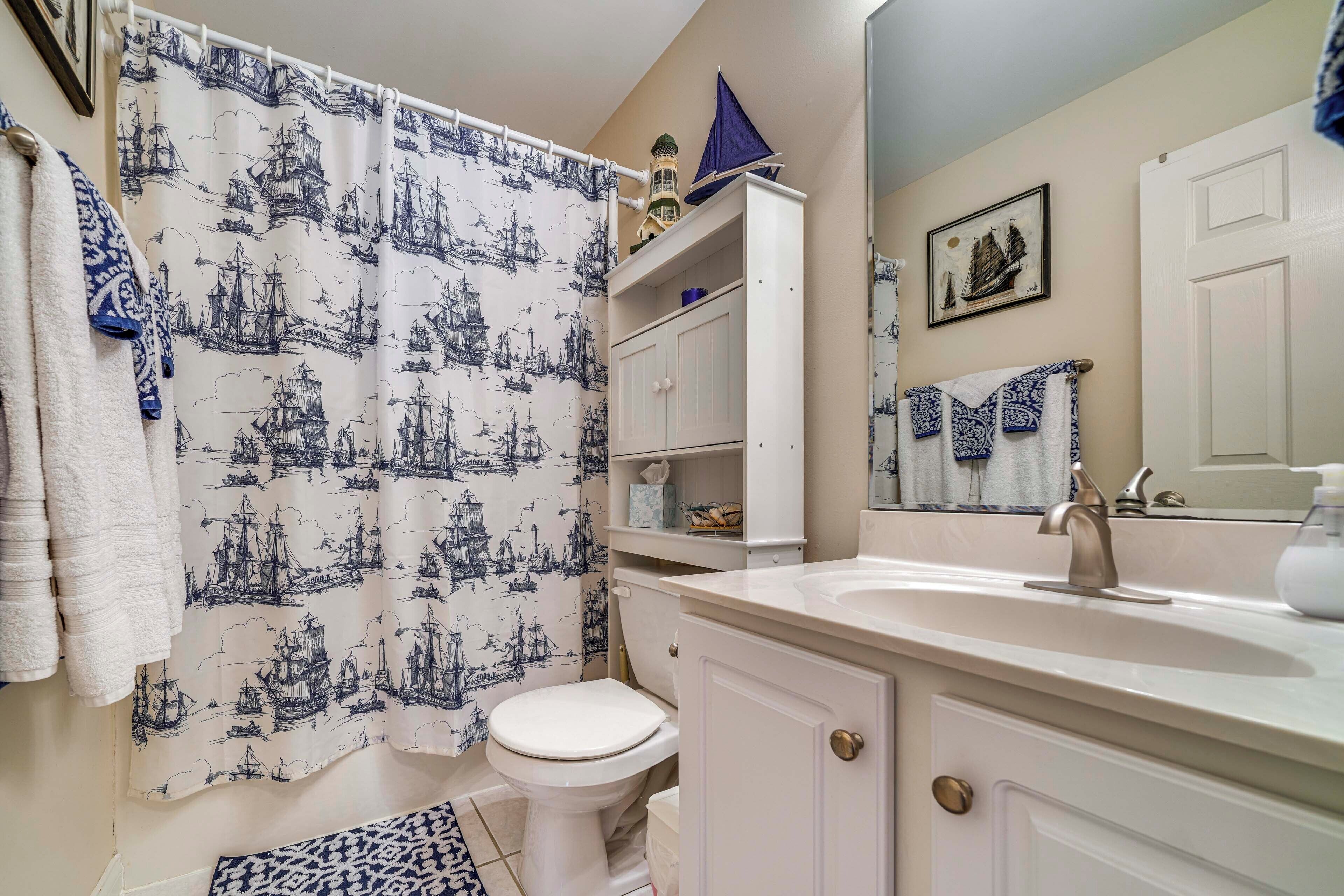 Un baño con cortina de ducha de temática náutica, un inodoro blanco, un lavabo con espejo y un estante con artículos de aseo.