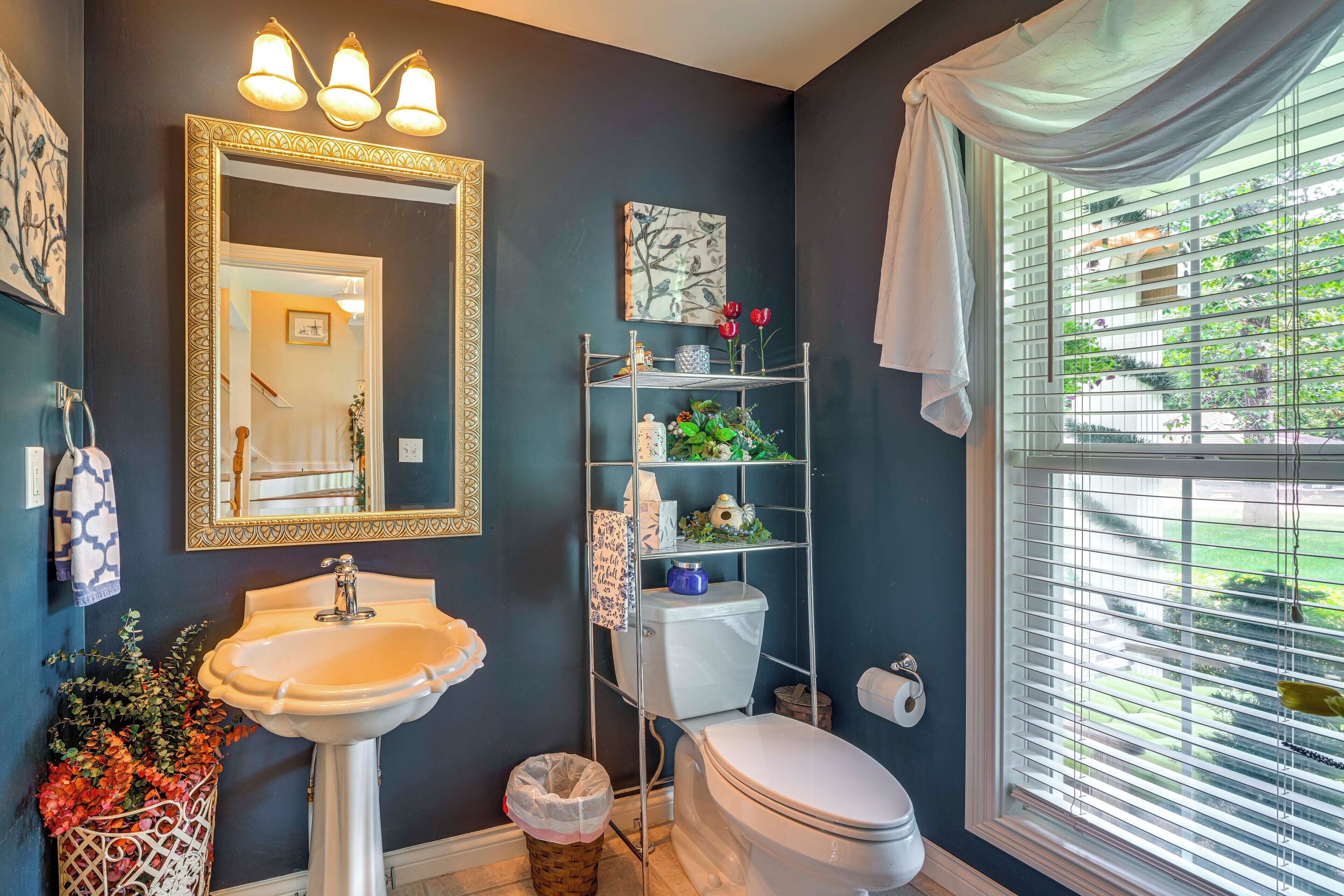 A bathroom with a pedestal sink, a toilet, a mirror with a decorative frame, a window with blinds, and a shelf with toiletries.