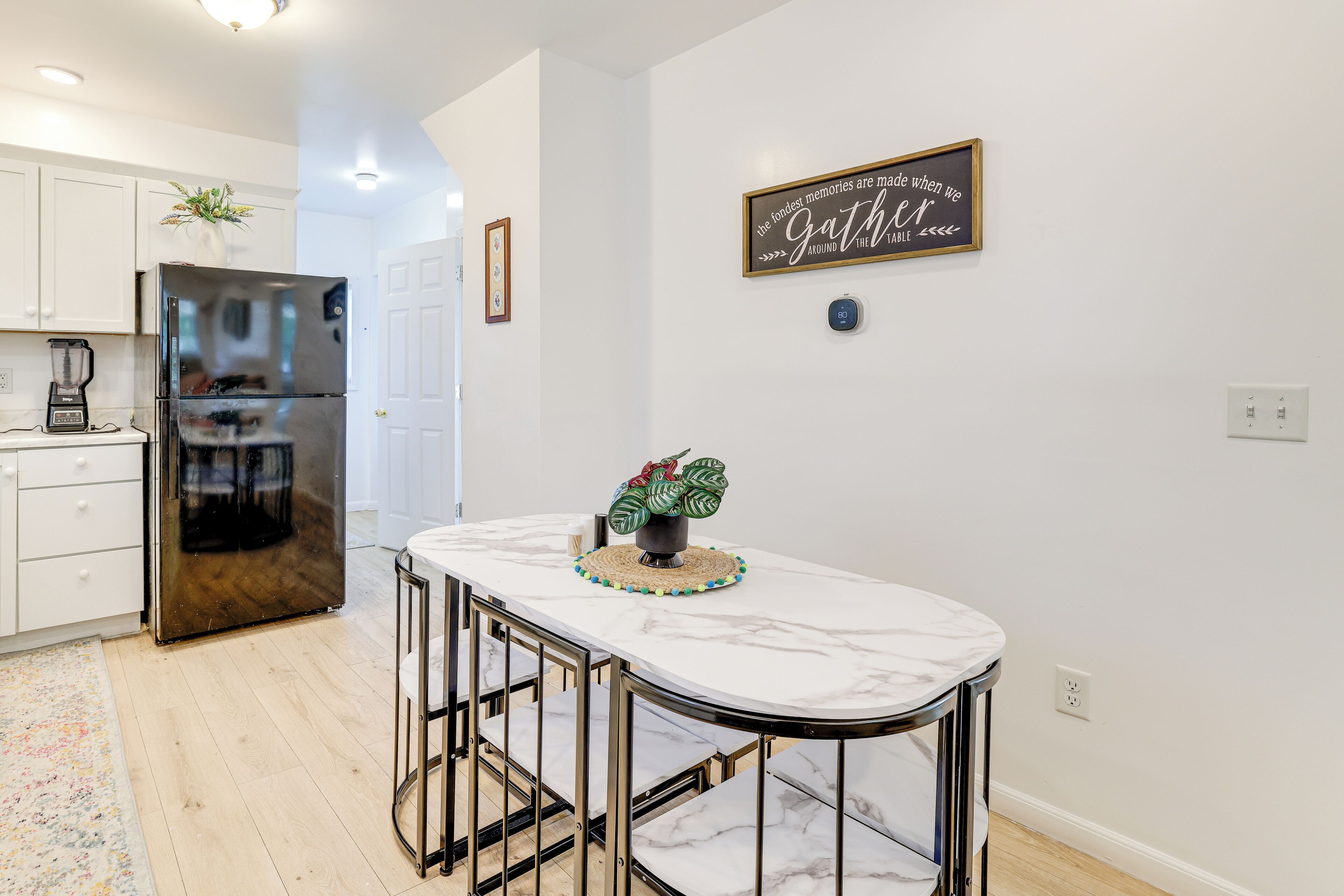 A modern kitchen with white cabinets, a stainless steel refrigerator, a marble island with bar stools, and a decorative plant.