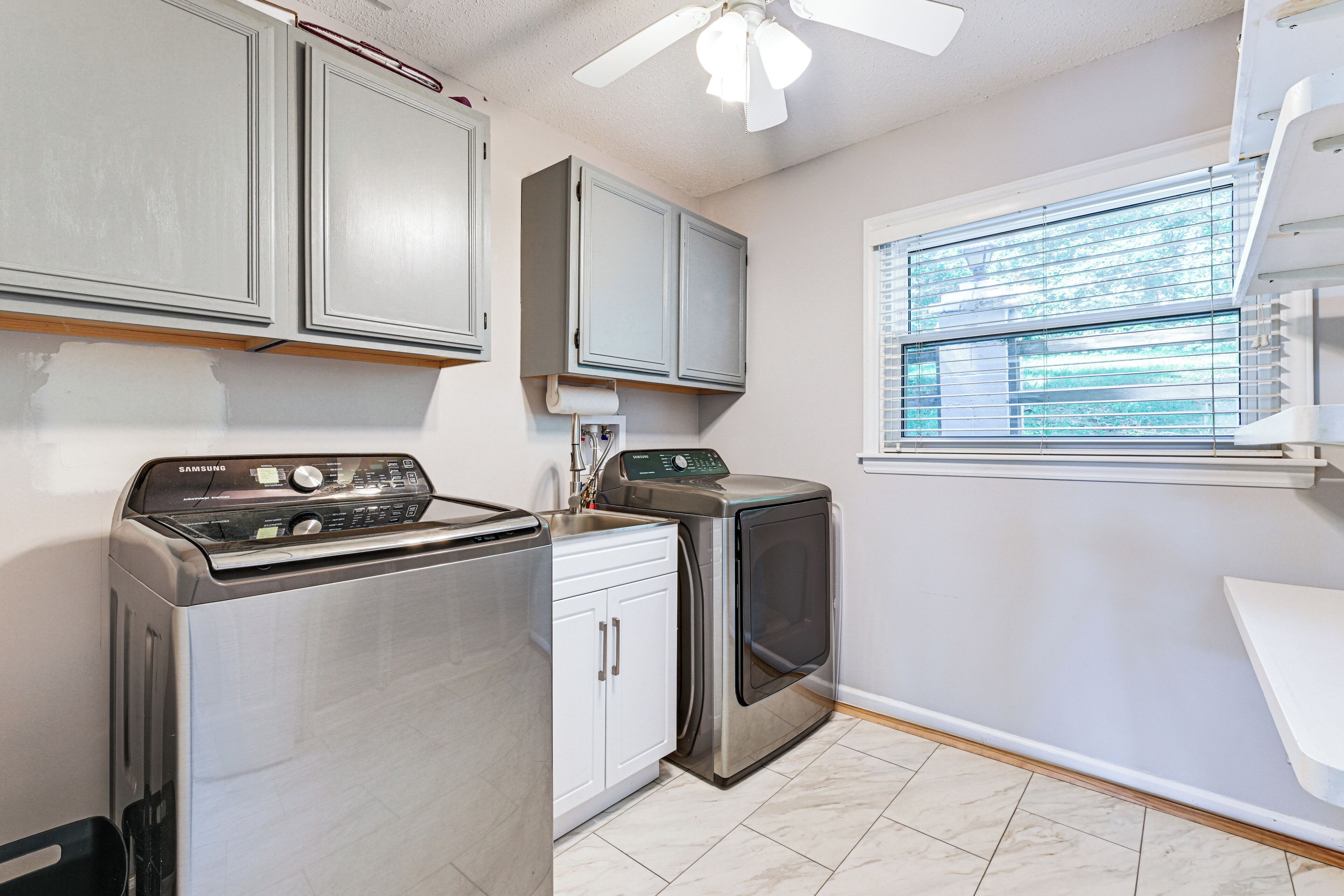 A compact kitchen with a stainless steel washer and dryer, white cabinets, and a window with blinds.