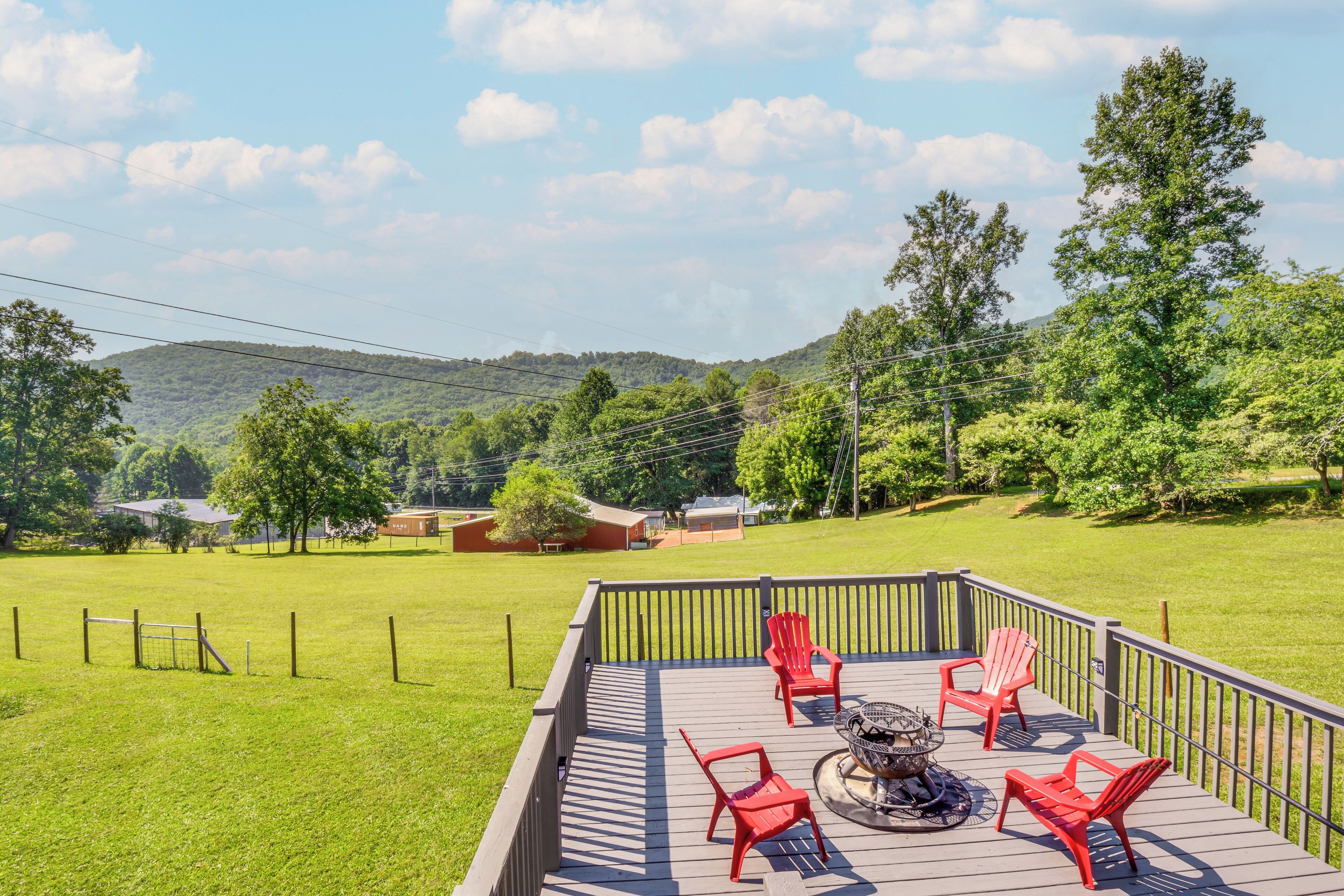 A deck with red chairs and a fire pit, overlooking a grassy area with trees and a distant hill.