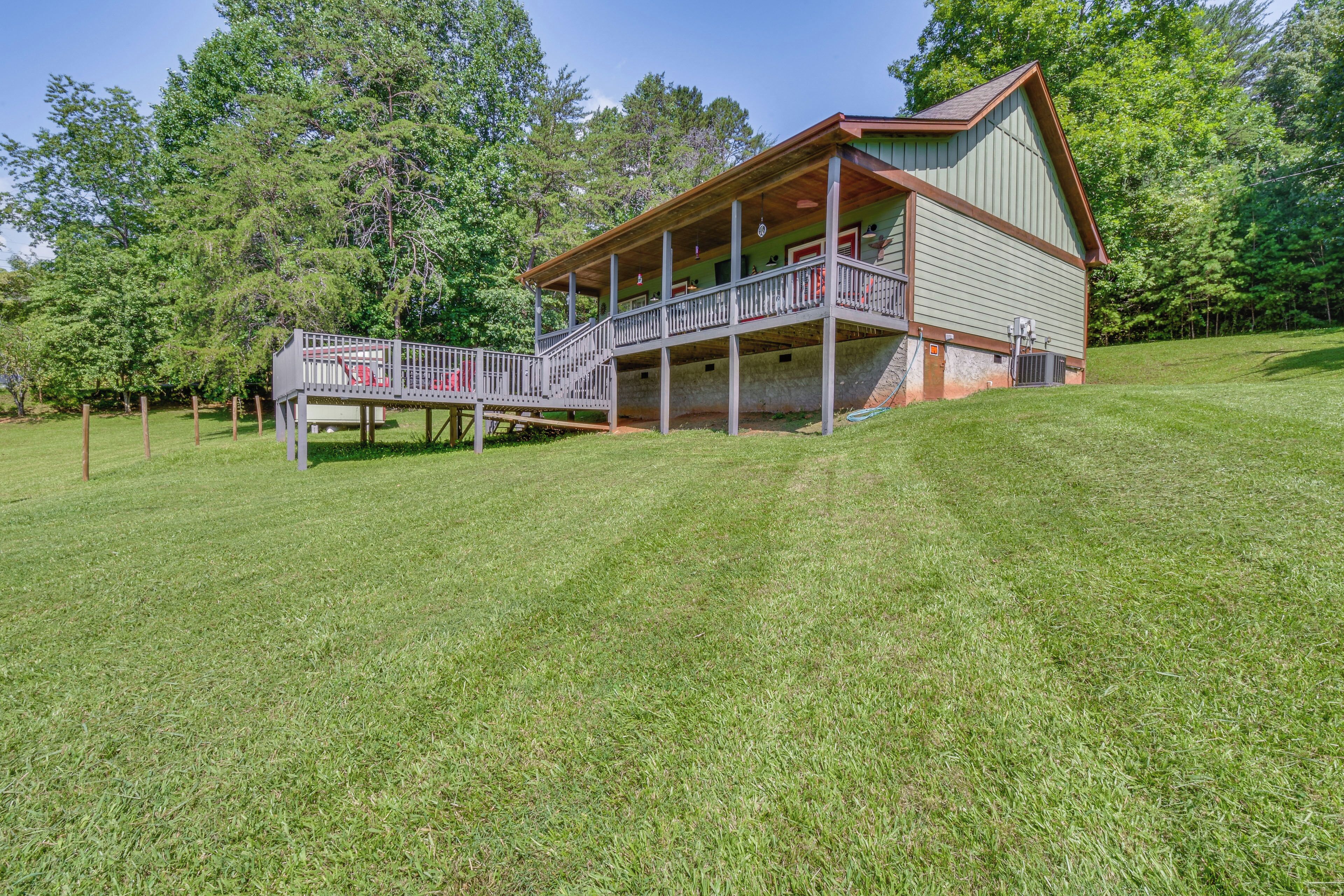 A two-story wooden cabin with a porch and a small deck, surrounded by a grassy yard and trees.