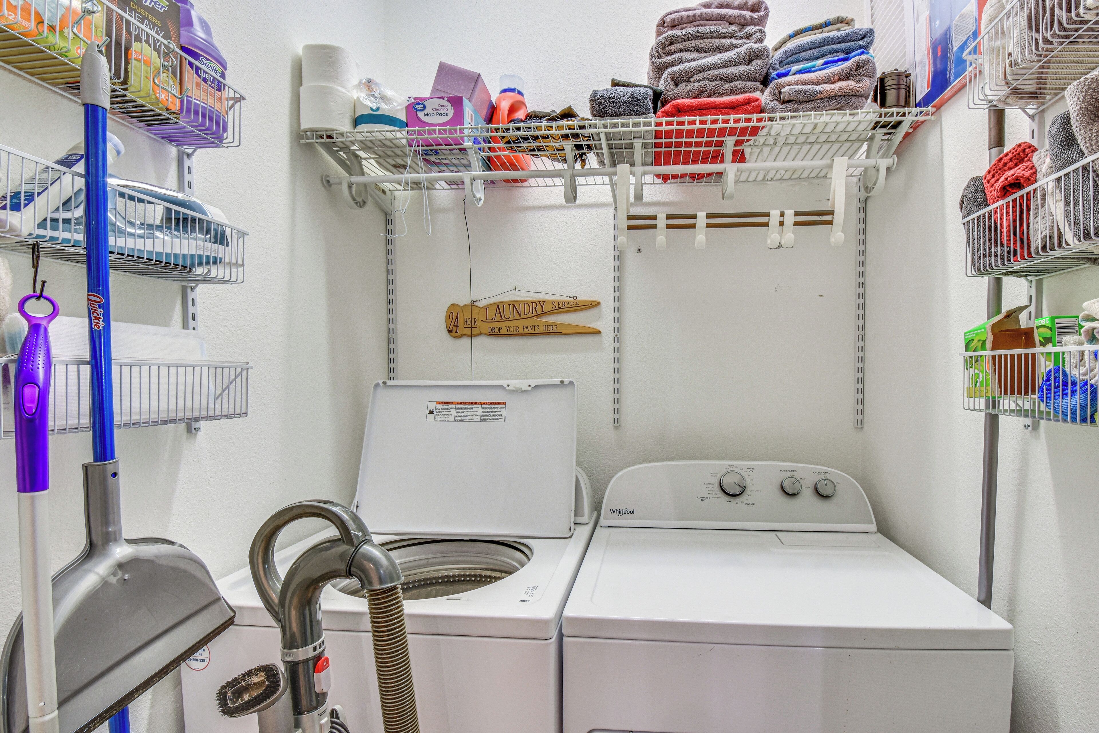 A laundry area with a front-loading washer and dryer, shelves with folded towels, and a sign indicating a 24-hour laundry service.