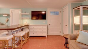Un espacio habitable compacto con zona de comedor que incluye una larga mesa de madera y taburetes blancos acolchados, un armario blanco empotrado con cajones, un televisor de pantalla plana montado en la pared y una pequeña mesa redonda de madera en la esquina.